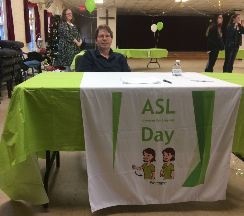 greeter at a table with a sign ASL Day, Welcome